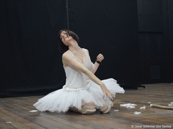 Pessoa ajoelhada num palco de madeira, com tutu branco, em pose expressiva de dança, rodeada por penas e um bastão.
