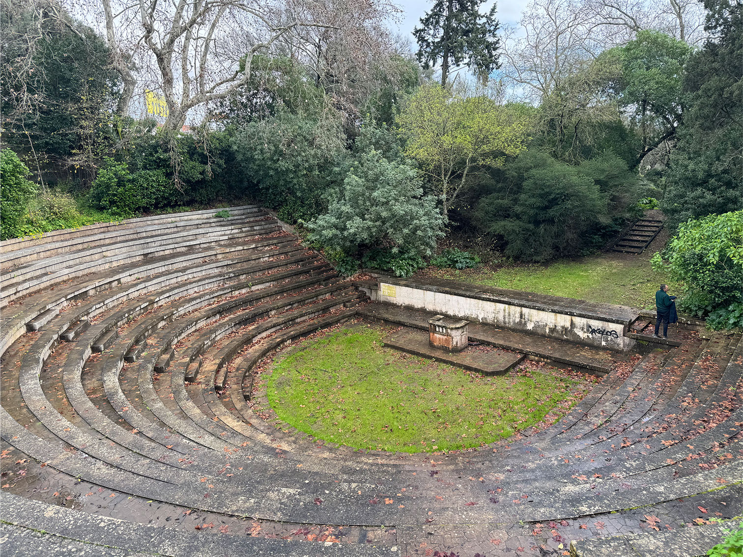 "An open-air stone amphitheater in a semicircle, with steps covered in dry leaves. At the center, a grassy area and a small stone structure. In the background, a low wall, dense vegetation, and a staircase. A person in a dark coat stands to the right near the forest, either observing or taking a photograph."