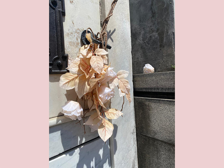 The image shows light-coloured artificial flowers hanging from a black metal fixture attached to a light stone wall, illuminated by sunlight.